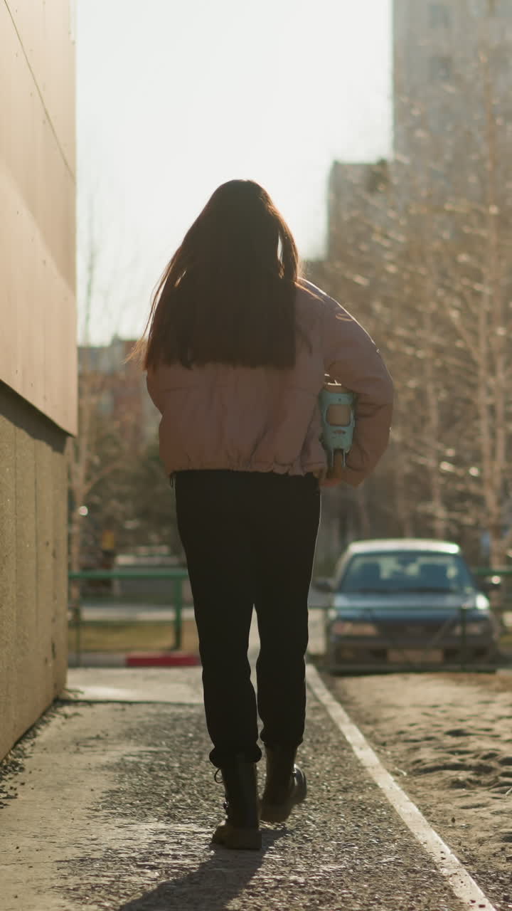 fotografía trasera de una chica con una chaqueta de melocotón y pantalones negros, sosteniendo una patineta turquesa, caminando por un camino iluminado por el sol al lado de un edificio, con árboles, coches y un apartamento en el fondo