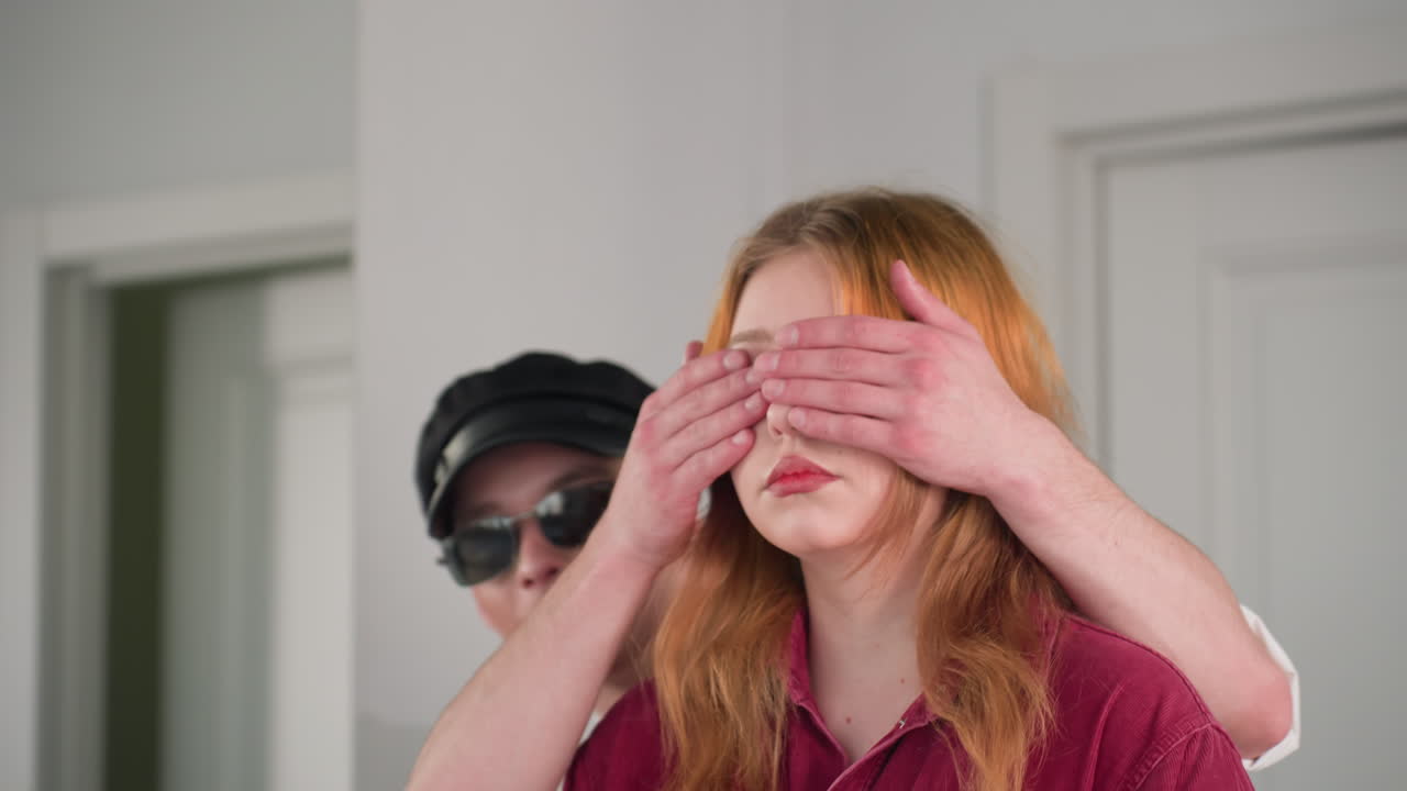 Close up of young lady in red shirt standing in front of door, looking to side with calm expression as unseen friend moves behind her to gently cover her eyes, beginning a playful interaction indoors