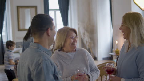 two happy middle aged women and man standing and talking at family party while holding glasses and having fun