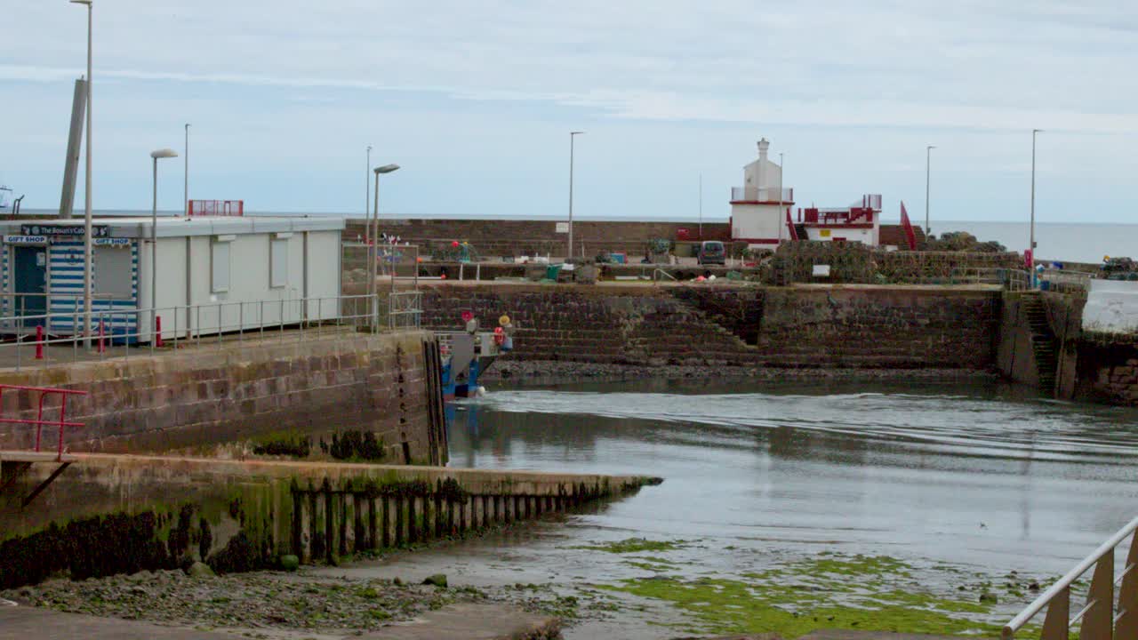 Blue fishing vessel enters stone harbor lock, overcast daylight, static wide shot, coastal environment