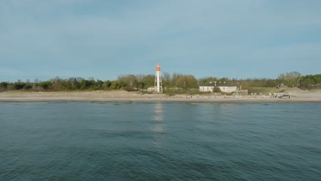 Aerial-establishing-view-of-white-colored-Pape-lighthouse,-Baltic-sea-coastline,-Latvia,-white-sand-beach,-large-waves-crashing,-sunny-day-with-clouds,-wide-drone-shot-moving-forward,-ascending