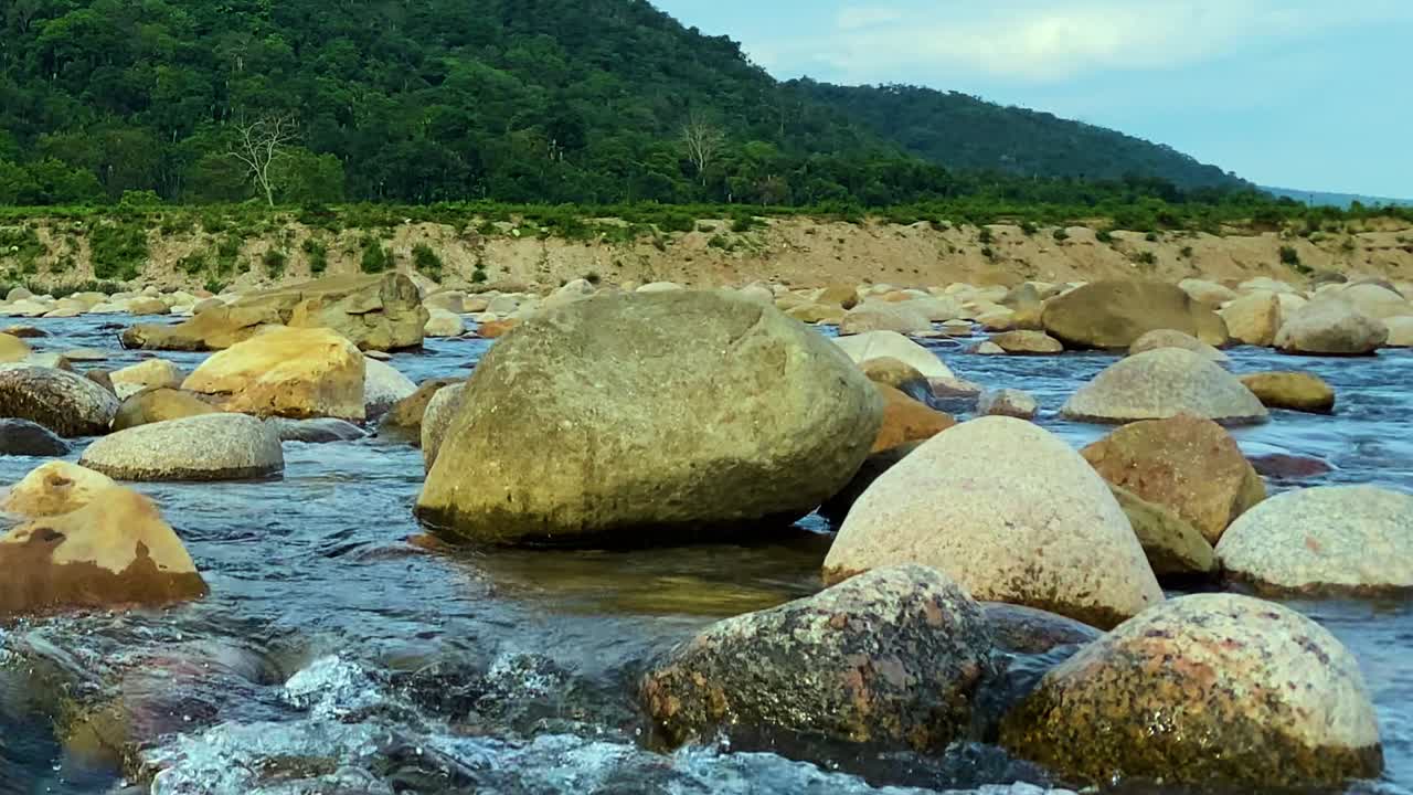 Low-angle view of a clear mountain stream rushing past large boulders with forested hills behind in Bichnakandi, Bangladesh, showing freshwater rapids and smooth stones