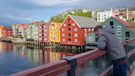 a man picks up a stone and playfully tosses it from a bridge into the nidelva river. behind him, colorful stilt houses reflect in the spring sunlight, filmed in trondheim, norway