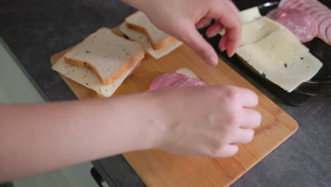 hands assembling sandwiches with sliced cheese and cold cuts on wooden cutting board in kitchen. several sandwiches partially made, ingredients like bread and meat placed neatly beside, in process of preparation