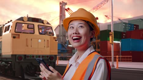 Close Up Side View Of Asian Female Engineer With Safety Helmet Using Smartphone And Looking Around With Freight Cargo Train At Port