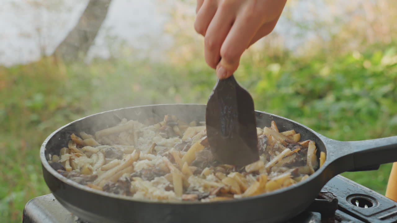 Padella da campeggio rivoltata e mescolata su fornello a gas portatile, mani che usano una spatola di metallo per mescolare patate e funghi, vapore che sale su uno sfondo verde lacustre, energico cuoco da campo che sta ultimando un piatto saporito.