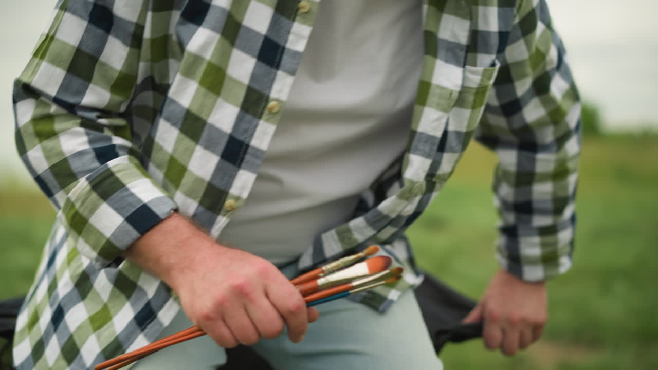 A close-up shot of an artist holding brushes, sitting on a chair in a grassy field, demonstrating a sitting pose. The checked shirt and outdoor setting emphasize the natural, creative atmosphere