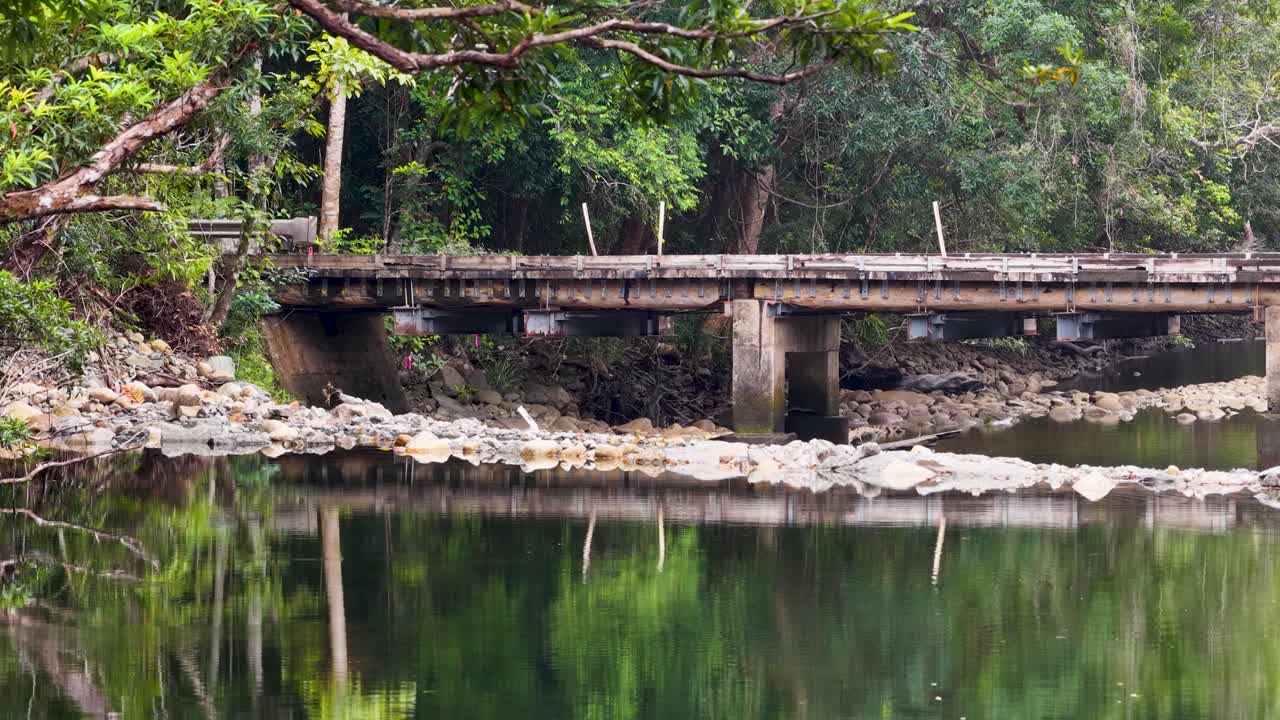 A white car crosses a rustic bridge over a calm river, surrounded by dense rainforest foliage