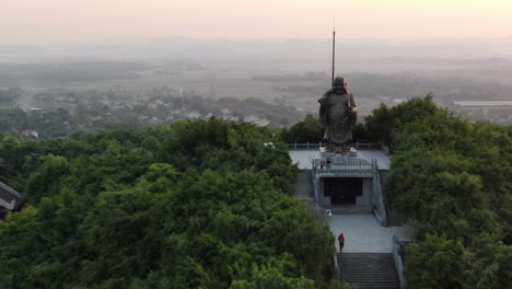 Drone-aerial-view-in-Vietnam-flying-towards-a-buddha-statue-surrounded-by-temples-on-a-green-tree-covered-valley-in-Ninh-Binh-at-sunset