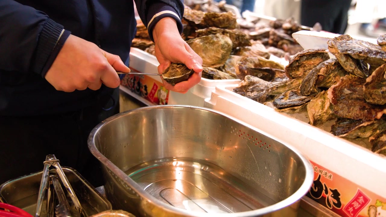A seafood vendor uses a knife to shuck fresh oysters at a busy outdoor market.