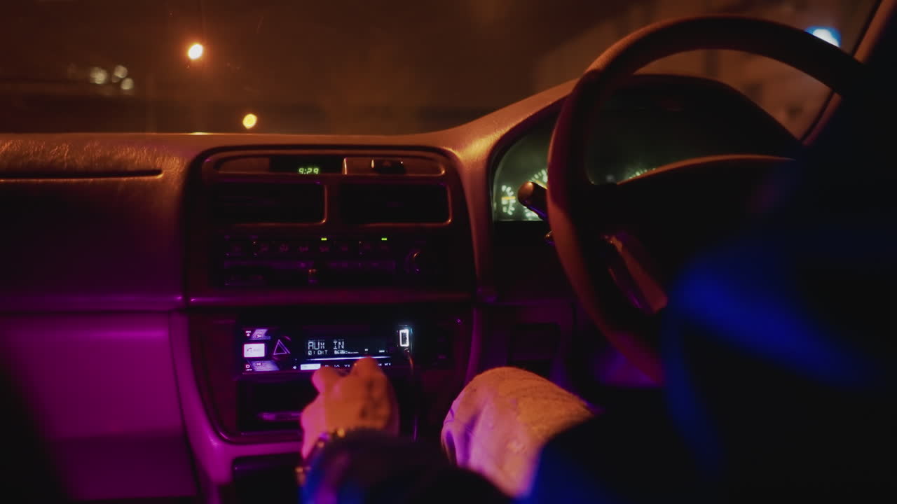 늦은 밤, 자동차 라디오 콘솔을 클로즈업하여 손으로 튜닝하는 모습, 네온 보라색과 파란색 실내 조명, 계기판 디스플레이가 켜져 있고, 손가락은 다이얼에, 미묘한 버튼 클릭 소리, 은밀하고 음악적인 분위기의 차 내부 장면