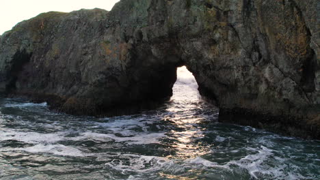 a close up of a large sea stack with a wave coming through an open tunnel