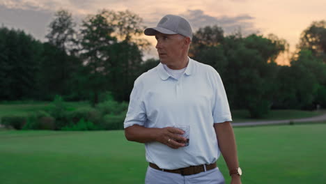 Pensioner-enjoy-rich-lifestyle-on-golf-field.-Old-man-posing-drink-glass-outside