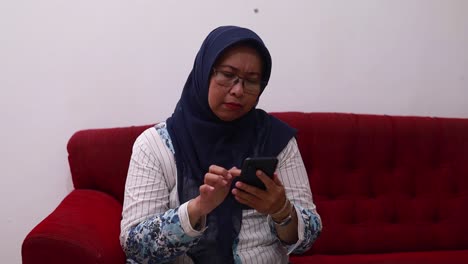 Veiled-asian-woman-sitting-on-the-sofa-at-home-while-using-a-cell-phone-with-unapy-face-expression