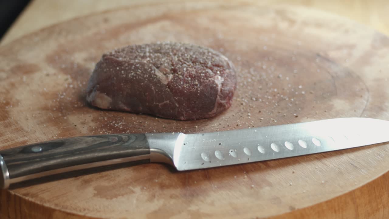 Ripresa slider al rallentatore di una bistecca che viene condita con sale e poi tamponata in cucina accanto a un affilato coltello da chef su un tagliere di legno spesso