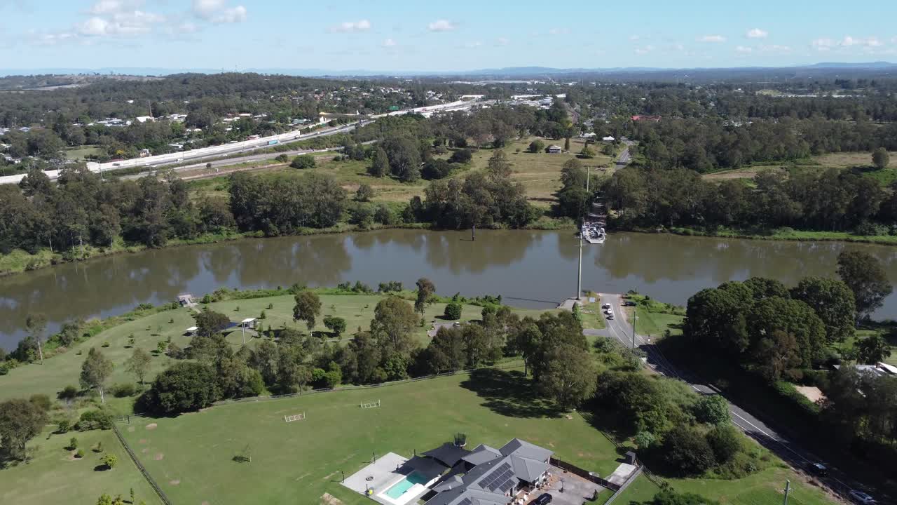 4K Aerial Video of a lush green rural suburb in Australia approaching a brown river and a ferry about to leave with cars on it