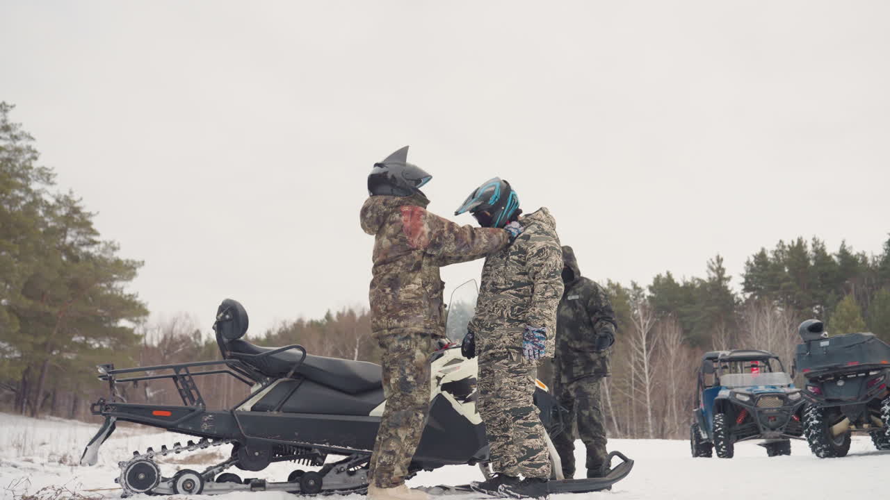 Two riders performing helmet check outdoors beside parked snowmobile, one adjusting visor and strap while crew looks on supportive mentorship, safetyfocused instruction and lastminute readiness create