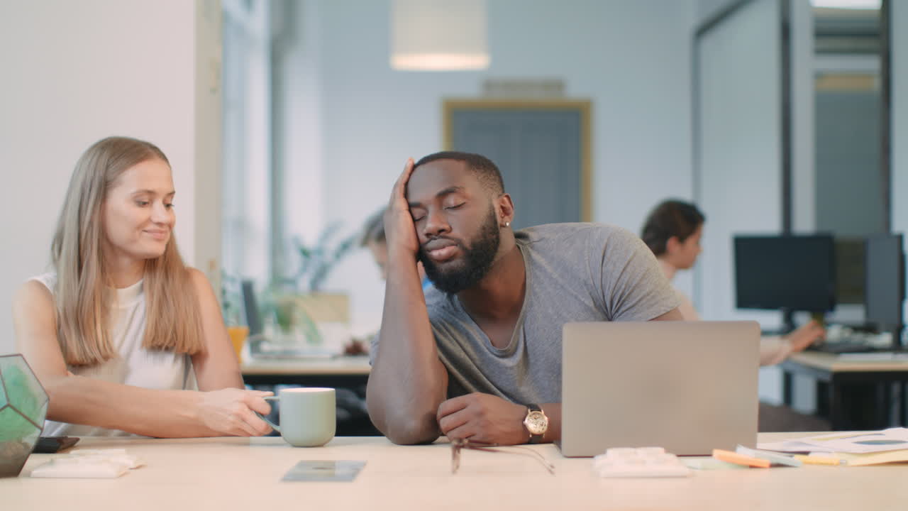 Tired guy falling asleep with laptop. Colleague giving coffee to sleepy man.