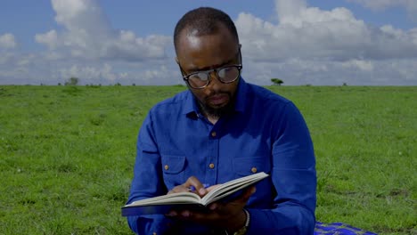 a black man sits in the grass and intently studies a book with pen in hand