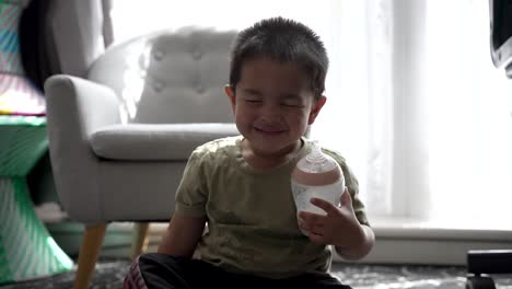 Adorable-Niño-De-18-Meses-Riendo-Con-Los-Ojos-Cerrados-Mientras-Sostiene-Una-Botella-De-Leche-Sentada-En-El-Suelo