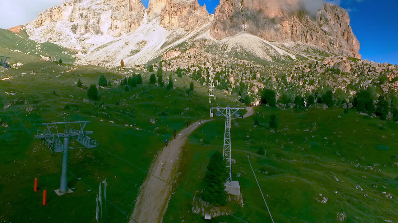 toma de drones del teleférico langkofel - sassolungo con excursionistas y ciclistas de montaña en el camino de abajo y el pico de langkofel en el fondo
