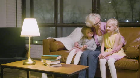 abuela abrazando a sus dos nietas sentadas en el sofá mientras habla en la sala de estar en casa 1