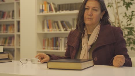 Moving-shot-of-an-angry-middle-aged-teacher-sitting-in-a-table-wearing-winter-coat-in-a-library-and-removing-glasses-by-looking-towards-the-camera