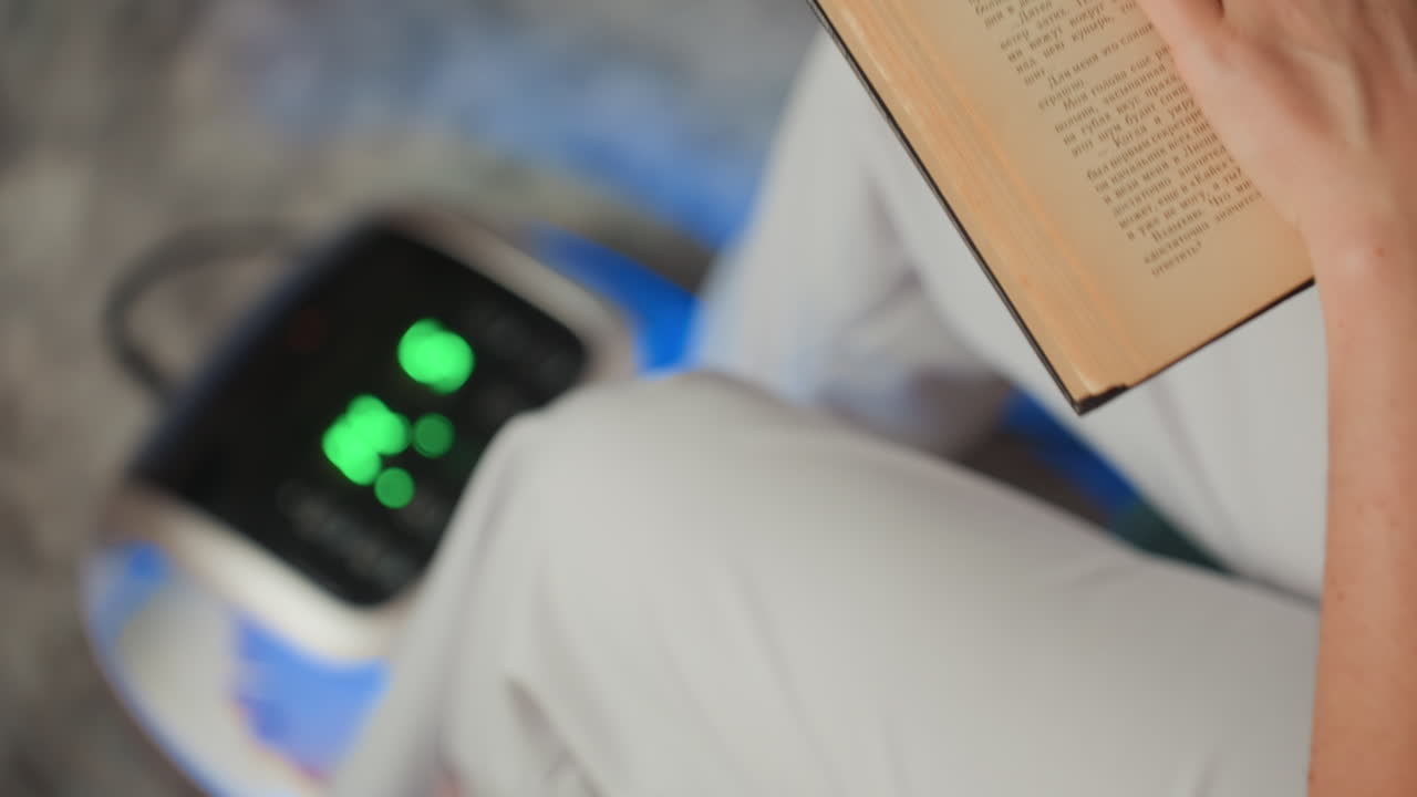 Paziente caucasica utilizza un dispositivo per i piedi a microcorrente mentre legge un libro tascabile, pantaloni bianchi e postura rilassata, display LED verde attivo, seduta in un salotto con focus sulla terapia plantare, atmosfera delicata