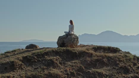 aerial view. flying the drone in a circle. a young woman in white clothes sits on a stone at the top of a hill against the background of the sea horizon.