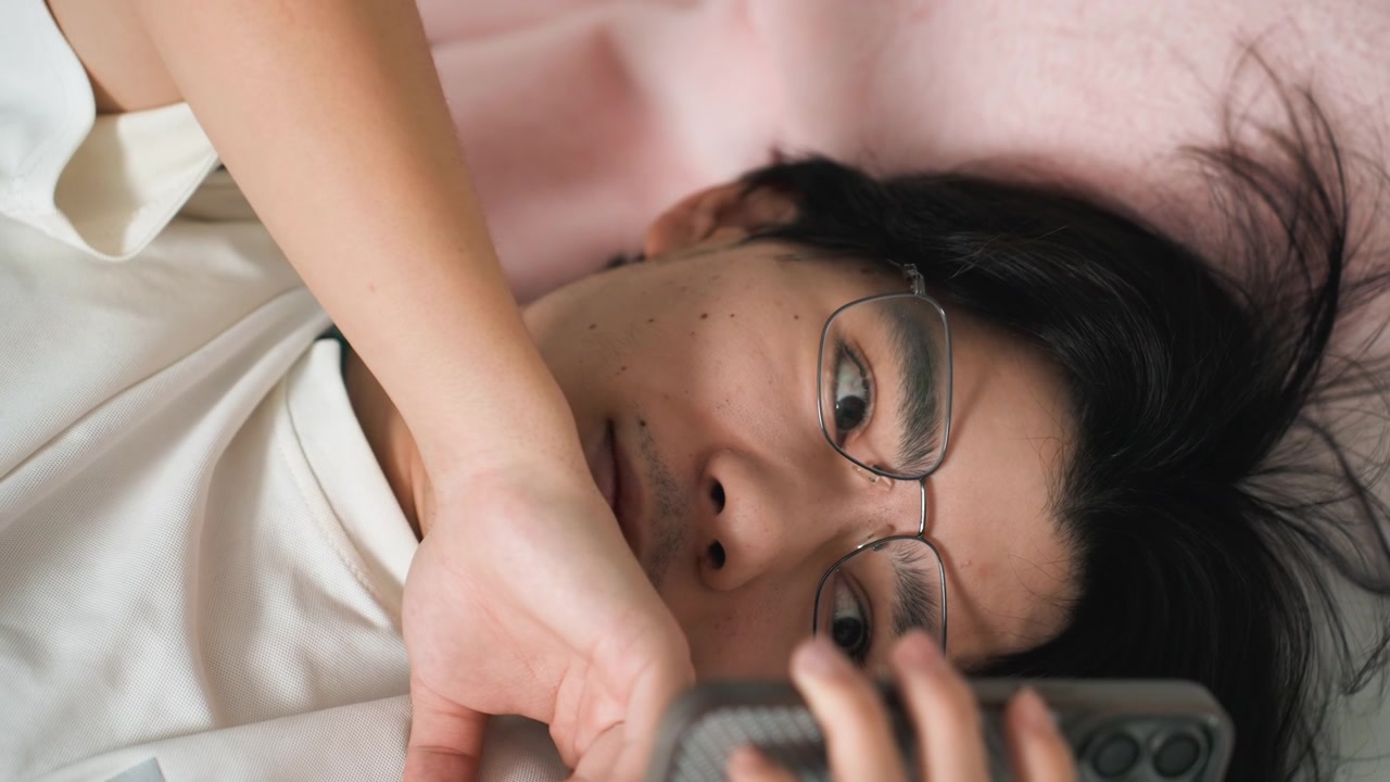 Scrittrice asiatica sveglia a letto che legge un messaggio sul telefono con gli occhiali, ritratto ravvicinato di espressione concentrata, sfondo di cuscini morbidi e luce soffusa, atmosfera domestica pensierosa e tranquilla.