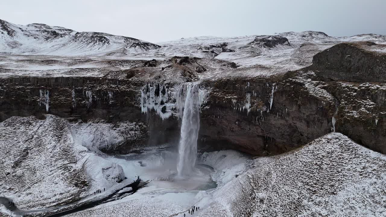 Aerial drone shot of Seljalandsfoss waterfall in Iceland during winter surrounded by snow-covered cliffs, frozen streams, rugged terrain, icy slopes, scenic views, people walking near base.