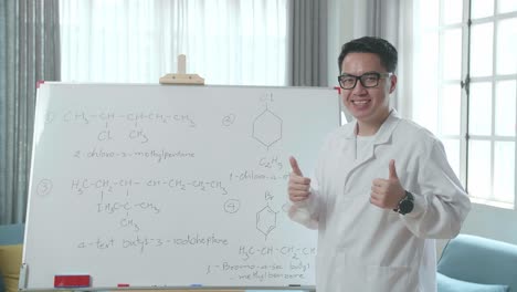 asian male teacher with glasses showing thumbs up gesture and smiling to camera while teaching chemist at home