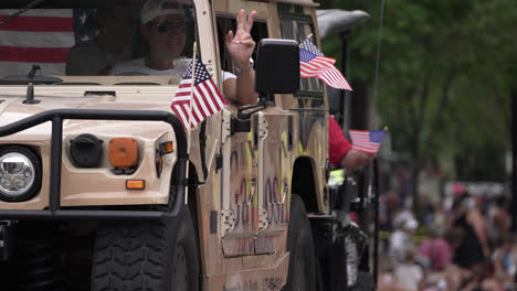 painted humvee drives in parade as people wave american flags with the stars and stripes outside of window to parade goers showcasing american patriotism