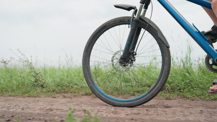 Ruota anteriore che gira su un sentiero sterrato, mountain bike blu con forcella ammortizzata, raggi e freno a disco che girano, scatto dal basso, sandali sui pedali, ciglio erboso, cielo nuvoloso, texture di ghiaia ruvida, attivo