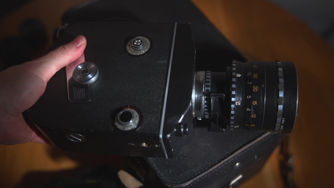 A close-up view of a vintage film camera with detailed metal controls and lens, partially held by a hand against a softly blurred warm indoor background