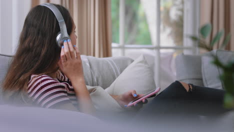 happy teenage girl listening to music wearing headphones relaxing on sofa at home texting with smartphone enjoying sharing lifestyle on social media