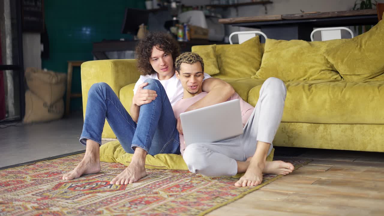 pareja gay romántica sentada en el suelo juntos, mirando la pantalla de la computadora portátil