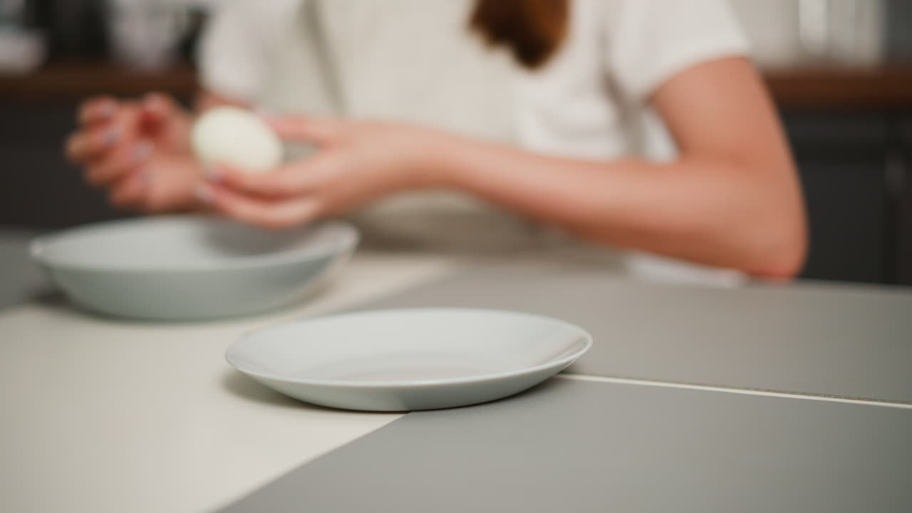 Slightly blurred view of woman collecting peeled egg from dish and returning it while using bowl to clean hand, with two plates on kitchen table and part of apron visible
