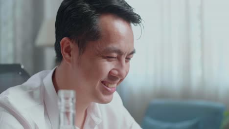 집에서 노트북으로 비디오 통화를 하는 동안 환호하고 술을 마시기 전에 글라스에 보드카를 부는 아시아 남자의 클로즈업