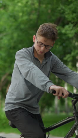 un niño joven con gafas se sienta en su bicicleta, conversando con su amigo con camiseta negra que está sentado, el niño se levantó y se movió hacia él, con vegetación en el fondo