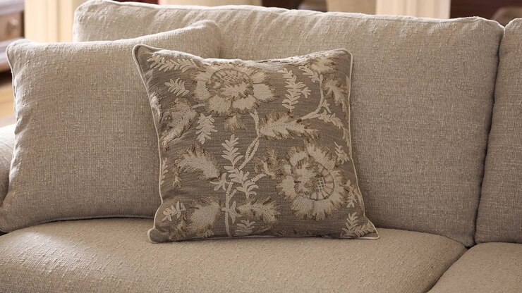 Lento zoom su un cuscino floreale beige chiaro appoggiato su un divano grigio, con la calda luce del sole che mette in risalto un soggiorno rustico arredato professionalmente.