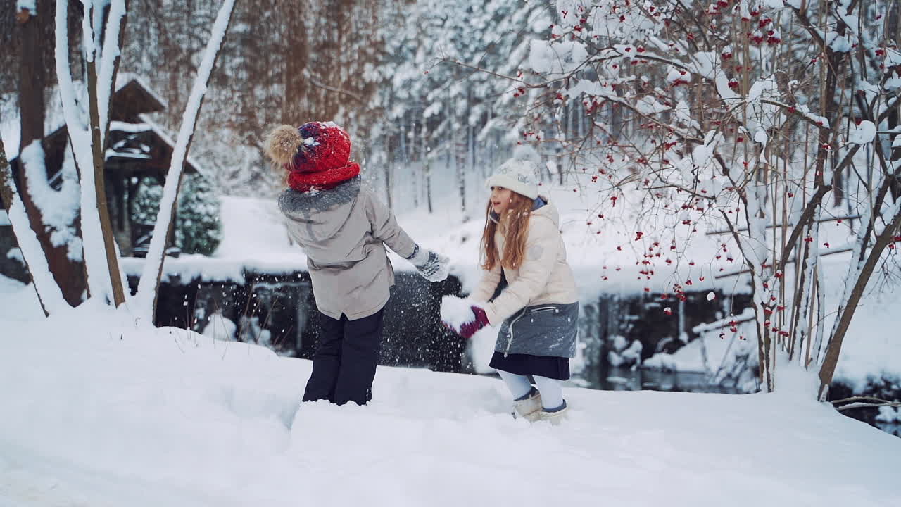 Happy children are playing with white snow together outdoors. Cheerful girls throw soft snow up over their heads in a beautiful forest in winter. Slow motion.