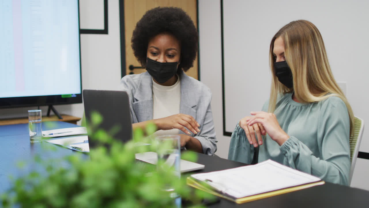 Two diverse businesswomen wearing face masks working together using laptop at desk in office
