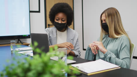 two diverse businesswomen wearing face masks working together using laptop at desk in office