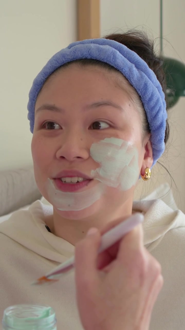 Video vertikal: Mencelupkan kuas, teman mengaplikasikan masker pada wanita dengan bando biru dalam rutinitas perawatan kulit di tempat tidur