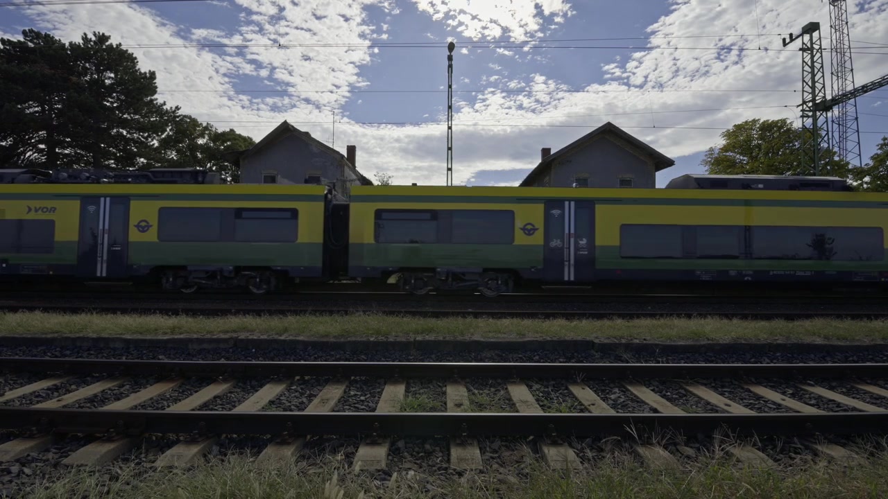 Un treno passeggeri elettrico passa accanto a vecchi edifici della stazione su binari ferroviari paralleli sotto le linee elettriche aeree in una giornata parzialmente nuvolosa