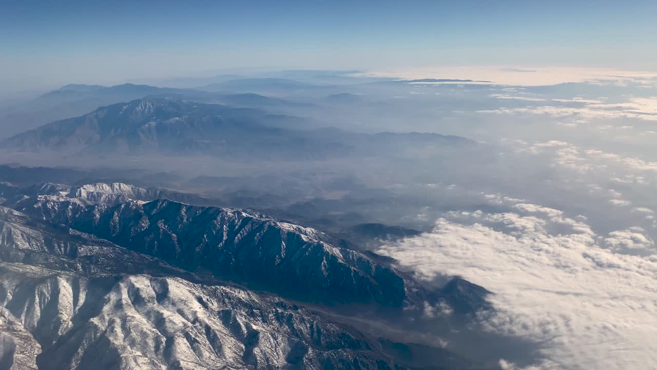 Vue aérienne depuis un avion volant entre Phoenix et Los Angeles, montrant des montagnes désertiques saupoudrées de neige, des crêtes superposées, des vallées brumeuses et des nuages bas sous une douce lumière du soleil