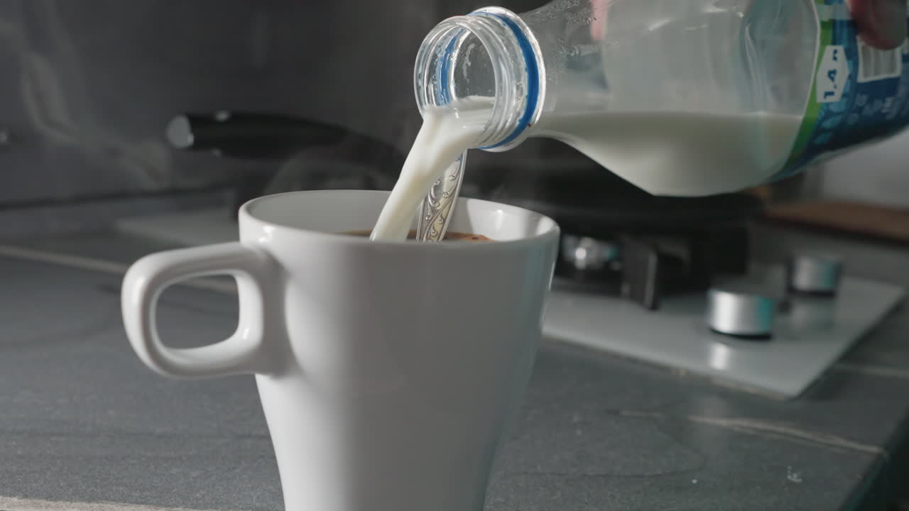Close-up of pouring milk into white mug. A person adds milk from bottle to cup, preparing a warm beverage. Fresh and clean kitchen atmosphere with soft light and modern kitchenware