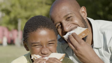 closeup of father and son eating sandwiches outside.