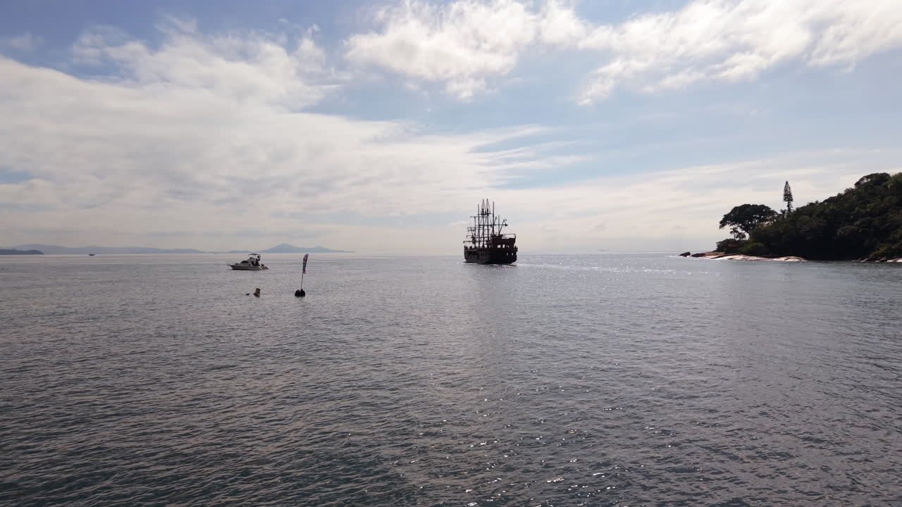 Tourist pirate ship sailing gracefully in the ocean near Balneario Camboriu, surrounded by a small island and boats, all under a cloudy sky on a tranquil day